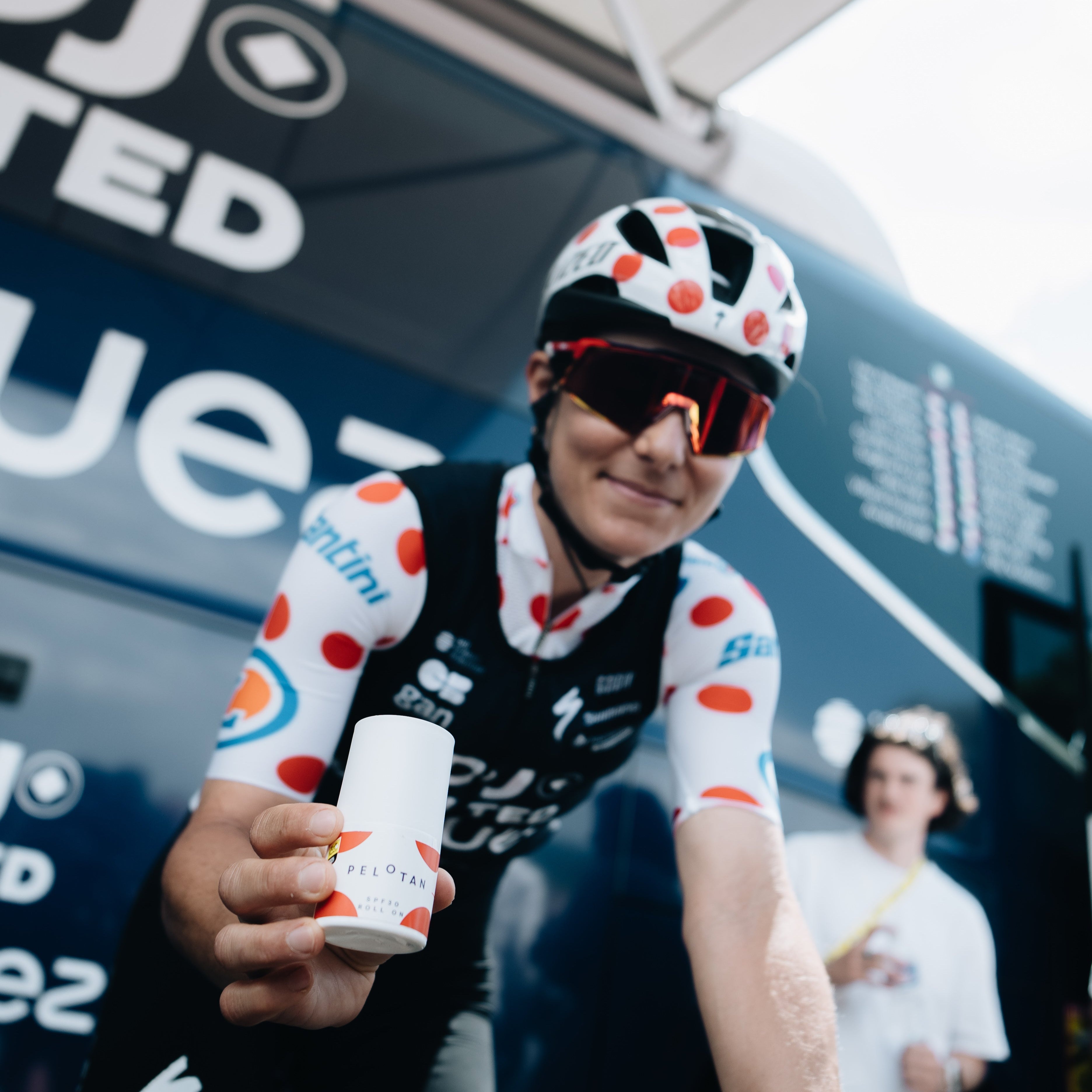 Cyclist in a polka dot jersey holding a Pelotan roll-on in front of a branded backdrop