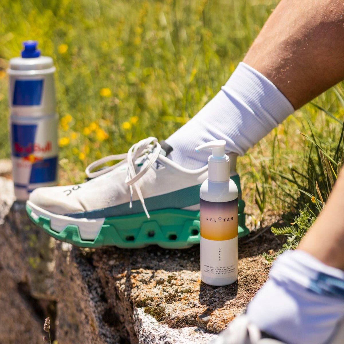 A bottle of Pelotan Apres recovery balm is on a rock, while an athlete sits on a grassy mountainside.