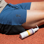 A runner is sat on a red asphalt track, with a bottle of Pelotan Après next to them.