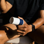 An athlete is holding a bottle of Pelotan 50ml Roll On performance sunscreen in their left hand.